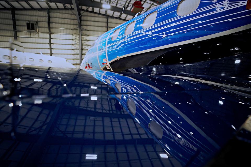 Nose of a business jet in deep blue metallic paint with fine white line graphics, mirrored in a glossy hangar floor.